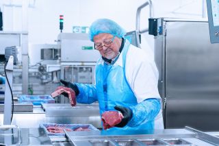 mannelijke werknemer van Dekeyzer-Ossaer in beschermende werkkledij aan het werk in de productieruimte, met stukken vlees in zijn handen